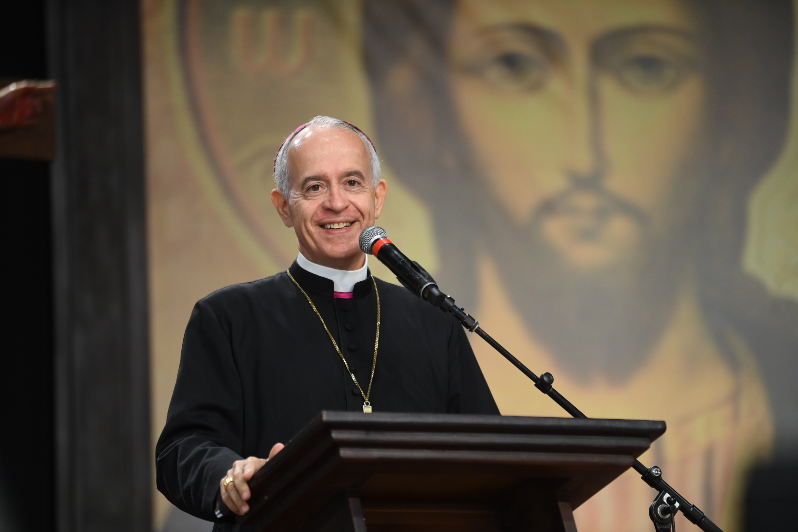 Um bispo católico sorridente, com veste preta, cruz peitoral e solidéu, fala ao microfone em um púlpito de madeira.