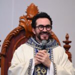 Um padre sorridente, com óculos de armação escura e barba, está sentado em uma grande cadeira de madeira trabalhada. Ele veste uma casula litúrgica clara com bordados em azul e ouro, segurando um microfone próximo ao peito com as duas mãos. Tema: "Vida nova ao sermos gerados em Maria"