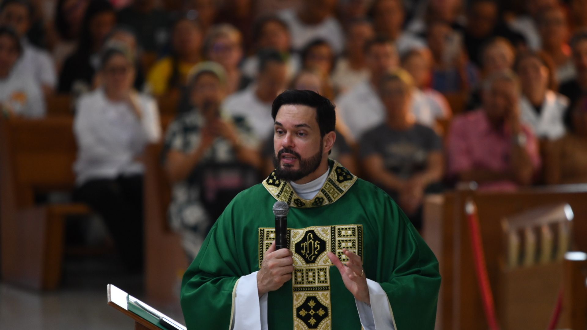 Padre de vestes verdes prega atentamente com o microfone na mão, gesticulando para enfatizar a sua mensagem.Ao fundo, avista-se a assembleia de fiéis sentada nos bancos da igreja, atenta à homilia.