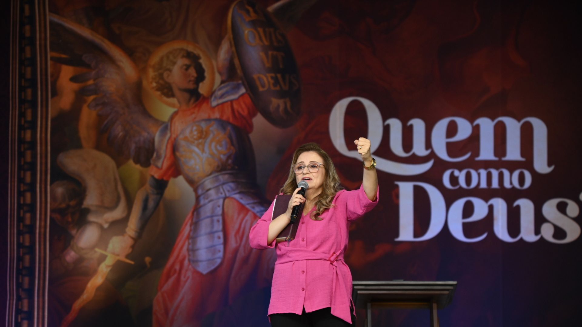 Mulher de camisa rosa e óculos fala ao microfone no palco, segurando um livro e erguendo o punho esquerdo num gesto firme.Atrás dela, vê-se a pintura do Arcanjo São Miguel e a frase em destaque "Quem como Deus".