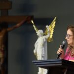 Uma mulher de cabelo castanho claro e óculos fala ao microfone num púlpito, vestindo uma camisa rosa. Ao fundo, desfocados, veem-se uma estátua do Arcanjo São Miguel de asas douradas e um crucifixo de madeira. Tema: "Houve uma batalha no Céu"
