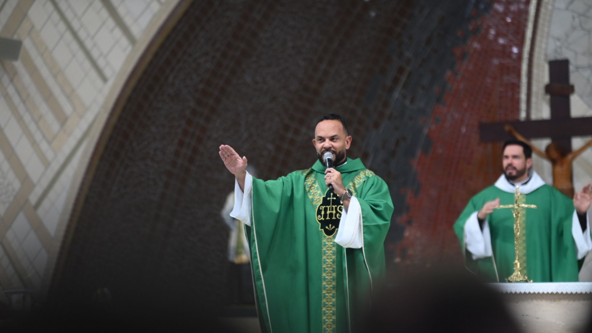 Padre com vestes verdes, de mão erguida a abençoar a assembleia enquanto segura o microfone. Tema: "Quem é Jesus para você?"