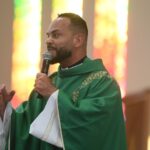 Padre vestindo casula verde e túnica branca, segurando o microfone com a mão esquerda enquanto gesticula com a direita durante a pregação. Tema: "Quem é Jesus para você?"