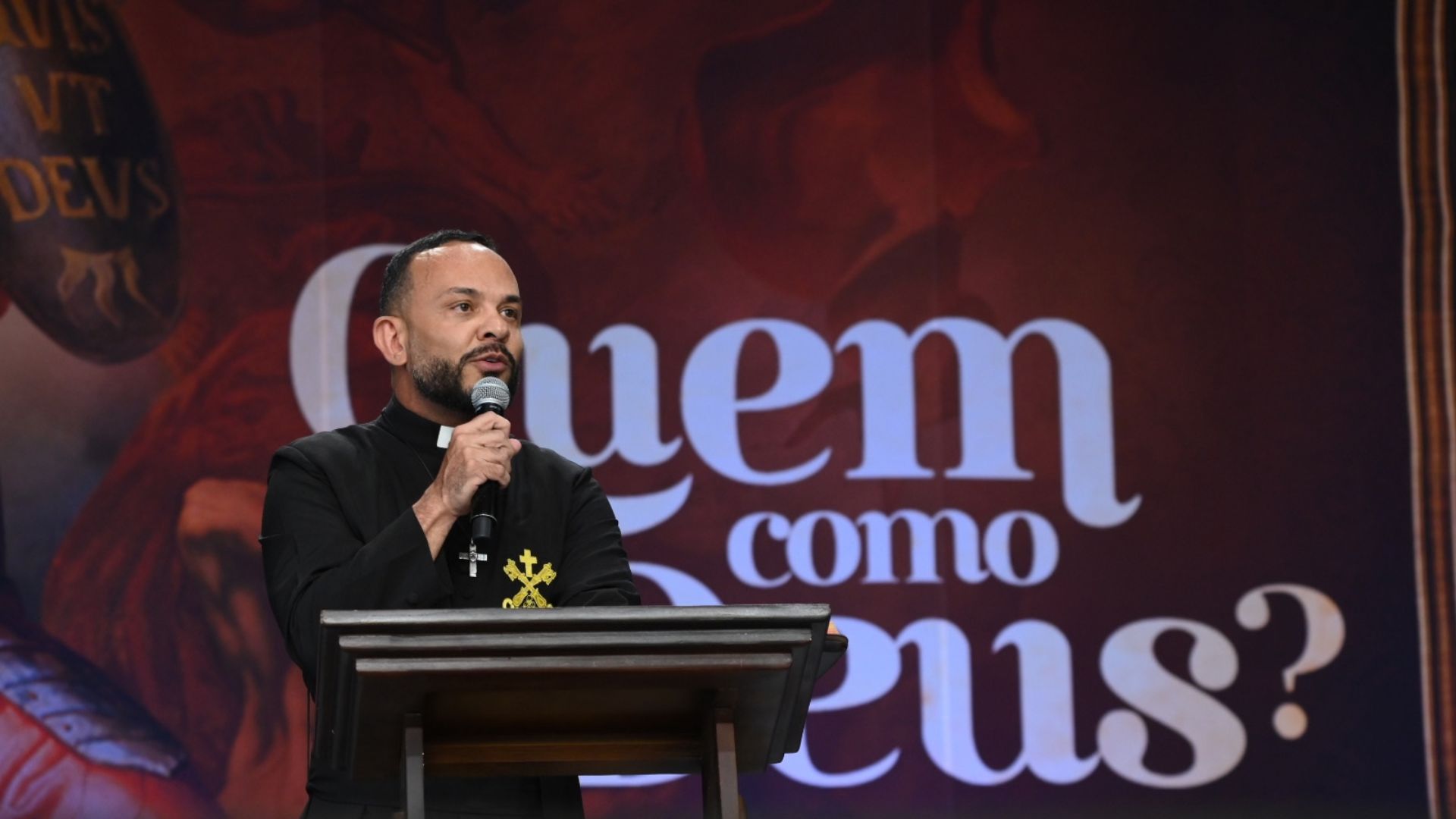 Padre de batina preta a falar ao microfone num púlpito de madeira.Ao fundo, destaca-se um painel em tons avermelhados com a frase emblemática "Quem como Deus?" escrita em letras brancas de grande dimensão. Tema: "São Miguel e a batalha espiritual"