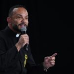 Padre de batina preta com colar clerical e um bordado dourado na vestimenta, segurando um microfone com a mão direita. Tema: "São Miguel e a batalha espiritual"