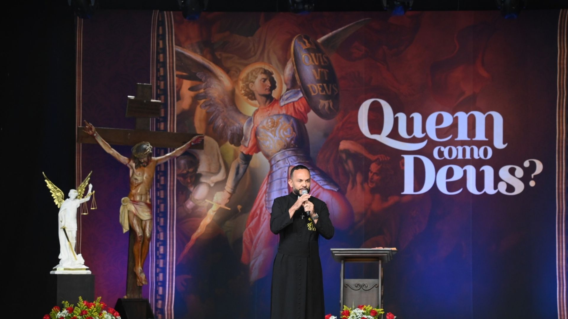 Um padre fala ao microfone num palco ao lado de um crucifixo, com um grande painel ao fundo ilustrado com São Miguel Arcanjo e a frase "Quem como Deus?". Tema: "São Miguel o Guardião da Igreja"