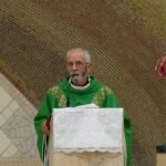 Padre de túnica verde fala ao microfone num púlpito. Ao fundo, destaca-se uma estátua de Nossa Senhora Auxiliadora