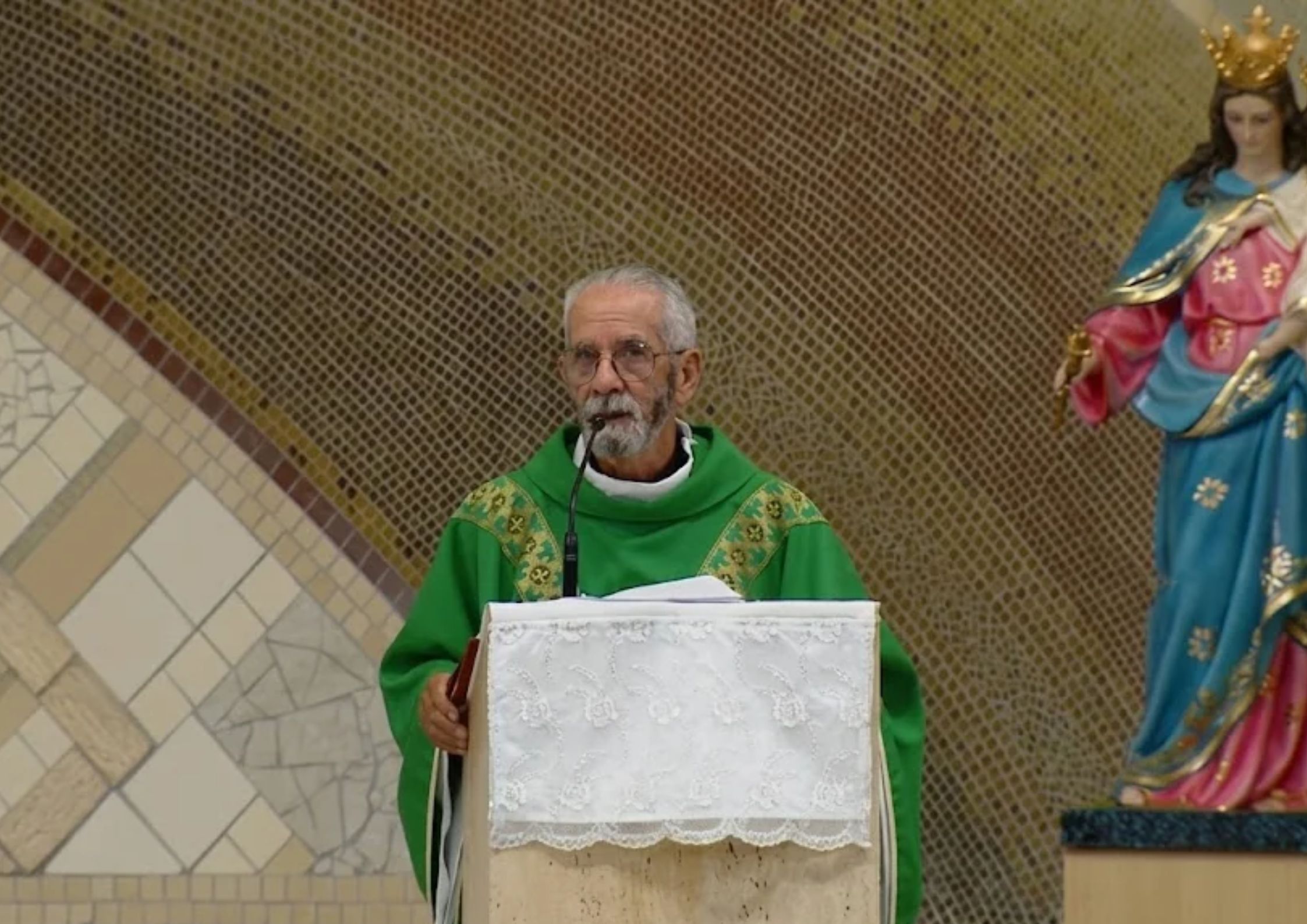 Padre de túnica verde fala ao microfone num púlpito. Ao fundo, destaca-se uma estátua de Nossa Senhora Auxiliadora
