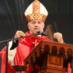 Bispo de vestes vermelhas e mitra ornamentada a discursar num púlpito de madeira trabalhada. Ele gesticula com as mãos enquanto fala para um microfone posicionado à sua frente. Tema: "A reconciliação oferecida na Cruz"