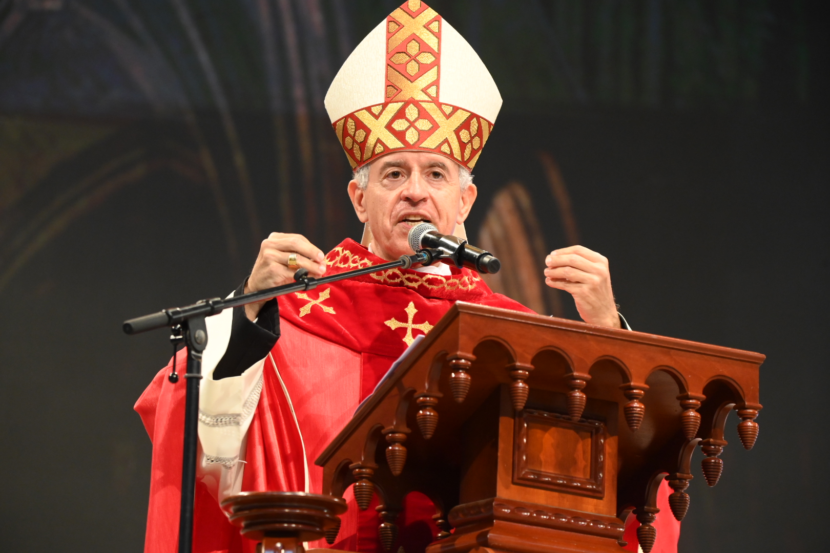 Bispo de vestes vermelhas e mitra ornamentada a discursar num púlpito de madeira trabalhada. Ele gesticula com as mãos enquanto fala para um microfone posicionado à sua frente. Tema: "A reconciliação oferecida na Cruz"