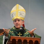 Bispo de veste verde e mitra ornamentada a discursar num púlpito de madeira trabalhada. Ele gesticula com as mãos enquanto fala para um microfone posicionado à sua frente. Tema: "Dar a Deus o que é de Deus"