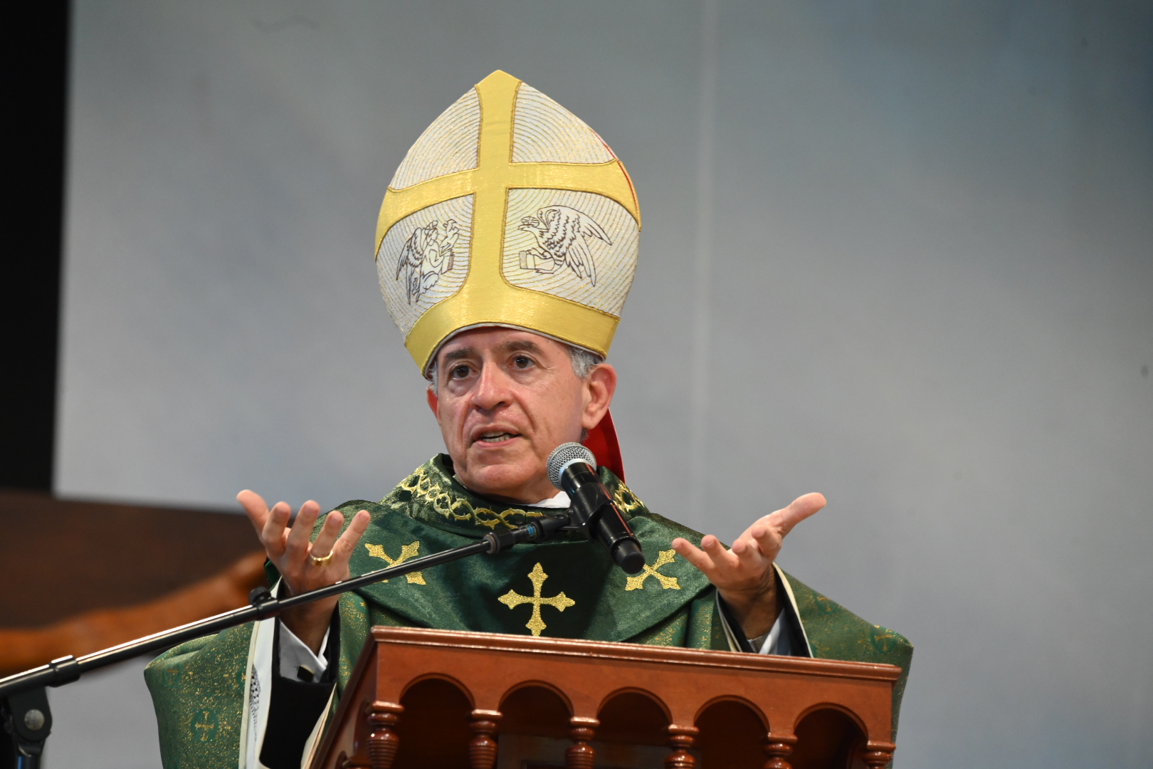 Bispo de veste verde e mitra ornamentada a discursar num púlpito de madeira trabalhada. Ele gesticula com as mãos enquanto fala para um microfone posicionado à sua frente. Tema: "Dar a Deus o que é de Deus"