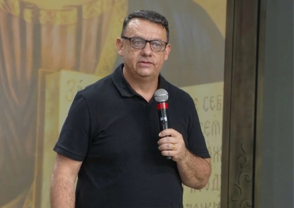 Um homem de óculos e camisa polo preta fala ao microfone enquanto segura o dispositivo com a mão direita.