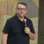 Um homem de óculos e camisa polo preta fala ao microfone enquanto segura o dispositivo com a mão direita.