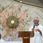 Um padre de barba grisalha prega ao microfone, de pé atrás de um púlpito de madeira com um crucifixo. Ele usa vestes brancas decoradas, com um grande ornamento de parede dourado e outro homem visíveis atrás. Tema: "Viver em estado de Graça"