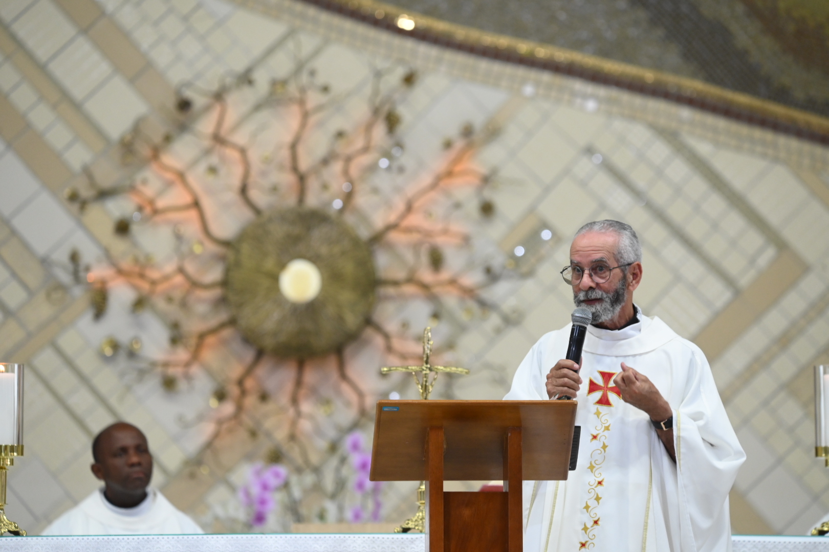 Um padre de barba grisalha prega ao microfone, de pé atrás de um púlpito de madeira com um crucifixo. Ele usa vestes brancas decoradas, com um grande ornamento de parede dourado e outro homem visíveis atrás. Tema: "Viver em estado de Graça"