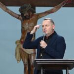 O homem de blazer azul fala ao microfone apontando para a cabeça, com um crucifixo ao fundo. Ele está de pé atrás de um púlpito de madeira, pregando em um ambiente de igreja. Tema: "O remédio para as nossas feridas"
