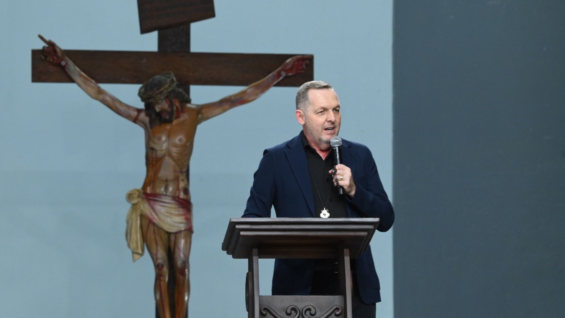 O homem de blazer azul fala ao microfone apontando para a cabeça, com um crucifixo ao fundo.Ele está de pé atrás de um púlpito de madeira, pregando em um ambiente de igreja. Tema: "O remédio para as nossas feridas"