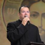 Padre veste batina preta e fala ao microfone enquanto segura a outra mão ao nível do peito. Ao fundo, destaca-se uma grande representação em tom dourado da imagem do rosto de Jesus Cristo. Tema: "Quando Deus diz não!"