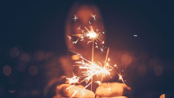 Mãos segurando velas faíscas brilhantes contra um fundo noturno desfocado, criando um ambiente festivo, mágico e cheio de luz radiante.