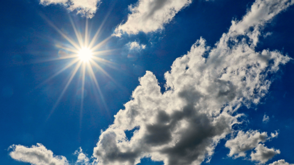 A cada novo dia são apresentadas uma nova beleza e descoberta Uma fotografia de grande angular do céu azul profundo, preenchido com várias nuvens brancas, fofas e cúmulos, com a bordas iluminadas pelo sol forte. O sol brilha intensamente no canto superior esquerdo, formando um poderoso flare estelar com múltiplos raios distintos e brilhantes que se estendem em forma de estrela. Um pequeno artefato de reflexão de lente circular verde pálido está visível à esquerda do sol, perto da borda da imagem. A composição captura o vasto céu azul, com a maior massa de nuvens ocupando o lado direito inferior e central. A luz solar intensa contrasta com as sombras suaves dentro das formações de nuvens, criando profundidade e textura. O céu é limpo e a luz é de meio-dia.