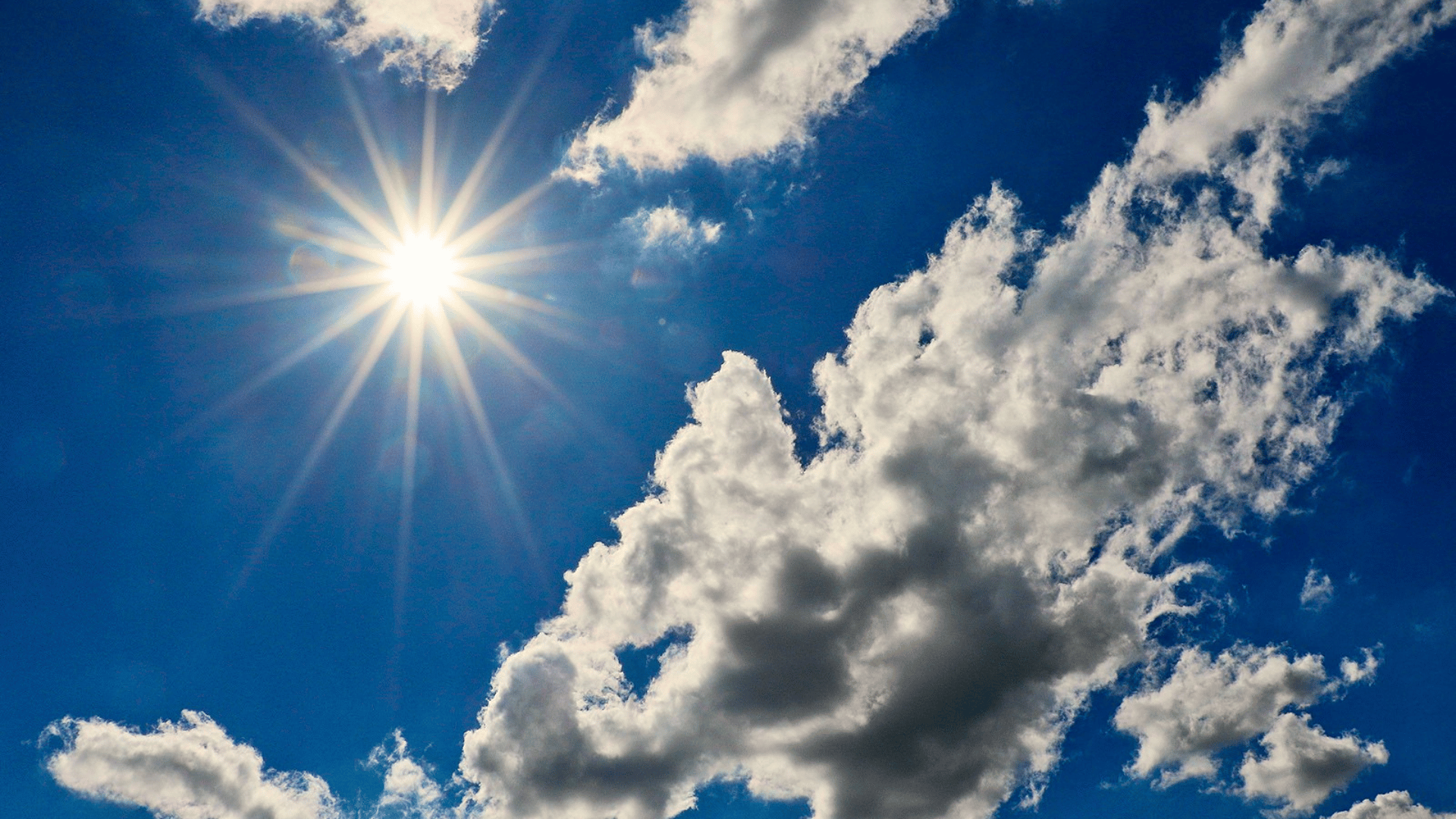 A cada novo dia são apresentadas uma nova beleza e descoberta Uma fotografia de grande angular do céu azul profundo, preenchido com várias nuvens brancas, fofas e cúmulos, com a bordas iluminadas pelo sol forte. O sol brilha intensamente no canto superior esquerdo, formando um poderoso flare estelar com múltiplos raios distintos e brilhantes que se estendem em forma de estrela. Um pequeno artefato de reflexão de lente circular verde pálido está visível à esquerda do sol, perto da borda da imagem. A composição captura o vasto céu azul, com a maior massa de nuvens ocupando o lado direito inferior e central. A luz solar intensa contrasta com as sombras suaves dentro das formações de nuvens, criando profundidade e textura. O céu é limpo e a luz é de meio-dia.