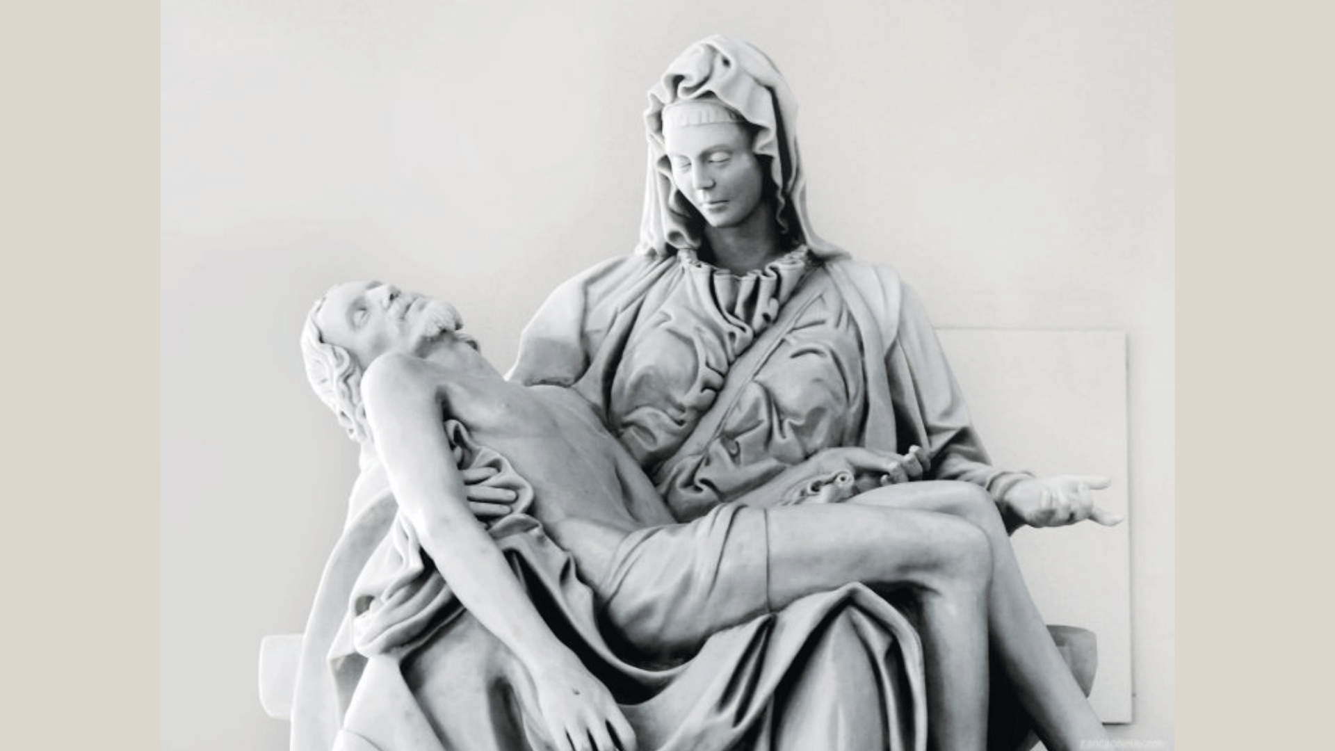 Escultura branca de Maria, com véu e expressão serena, segurando nos braços o corpo de Jesus Cristo após a crucificação. A cena retrata a Pietà, com Maria sentada e Jesus deitado sobre seu colo. Ao fundo, há uma parede clara e neutra.