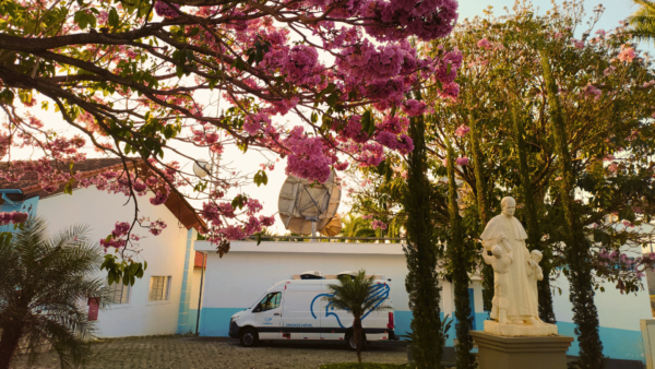 Fotografia panorâmica de uma área da Canção Nova, em Cachoeira Paulista, cercada por árvores e flores cor-de-rosa. No centro da imagem, há um prédio branco e azul com uma grande antena parabólica no telhado e uma van branca identificada como “Unidade Móvel”. Em primeiro plano, galhos floridos formam uma moldura natural sobre a cena, enquanto a luz do entardecer ilumina o ambiente. A imagem destaca a beleza da primavera, a missão de evangelização pelos meios de comunicação e o serviço da Canção Nova à comunidade.