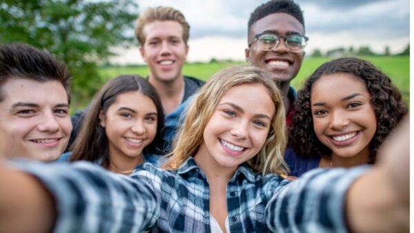 Jovens de diferente etnias, tirando uma selfie com o que parece ser uma tela de celular, eles sorriem, tendo ao fundo campos verdes, levemente desfocados