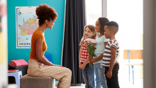 professor : sentada em um banco conversa com quatro crianças de pé, que estão entregando a ela um buquê de flores. A cena se passa em uma sala de aula e transmite um ambiente de carinho e gratidão.