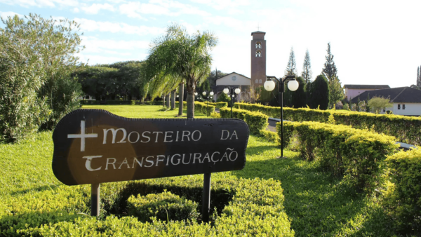 Vida monástica. Uma foto horizontal mostra uma placa de madeira preta e rústica na entrada de um mosteiro. A placa tem o formato de um escudo com uma cruz à esquerda. As letras brancas na placa dizem Mosteiro da Transfiguração.
