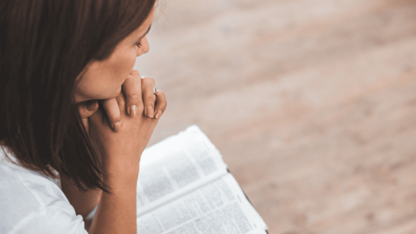 Espiritualidade : Foto de uma mulher com cabelos castanhos em um momento de devoção. Ela está de perfil, com as mãos juntas em prece sobre uma Bíblia aberta, com o fundo desfocado para destacar a cena de oração e reflexão.