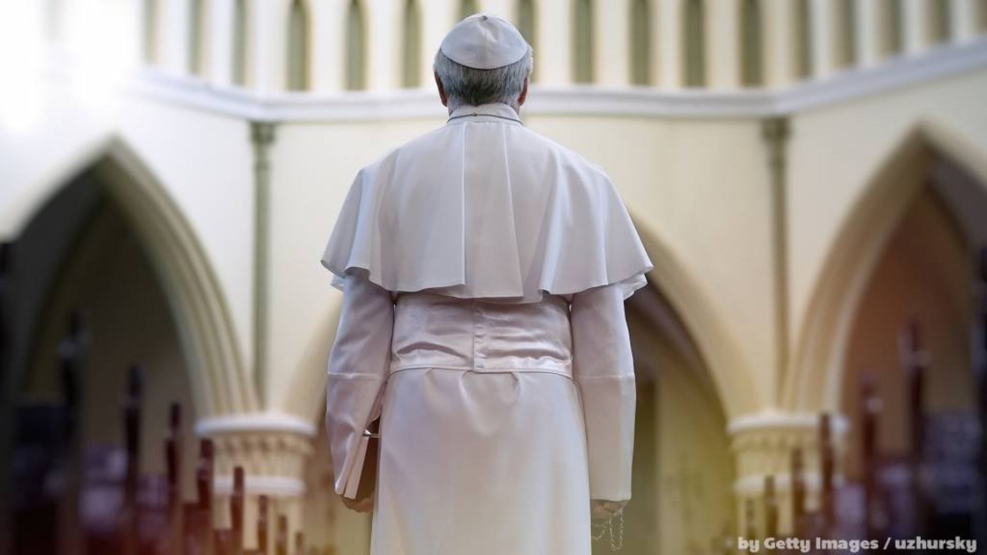 Papa de costas, em vestes brancas, em pé no corredor central de uma igreja clara, olhando para frente.