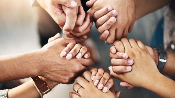 Várias mãos de diferentes tons de pele estão unidas em um círculo, segurando-se firmemente para simbolizar apoio, união e solidariedade.