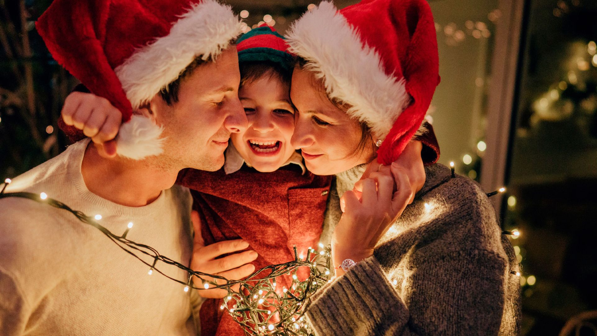 Uma família feliz de três pessoas usa chapéus de Papai Noel, abraçados e envoltos em luzes de Natal brilhantes e quentes.