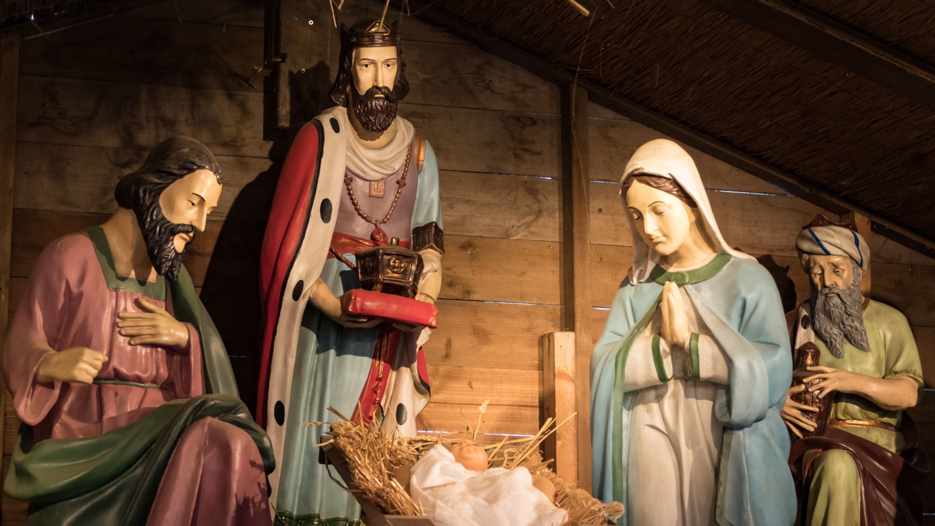 Uma cena detalhada de Natal com estátuas de São José, um Rei Mago, a Virgem Maria e outro Rei Mago ao redor do Menino Jesus em uma manjedoura de madeira.