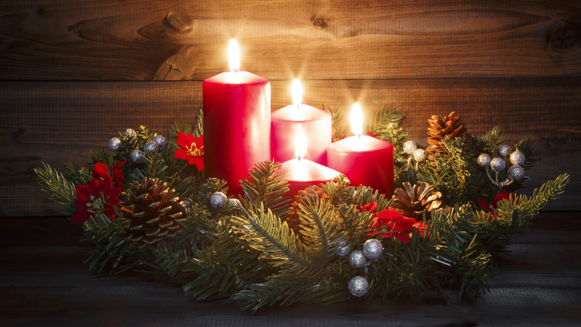 Quatro velas vermelhas acesas em uma guirlanda de Natal, iluminando pinhas e folhagens em um fundo rústico de madeira escura.