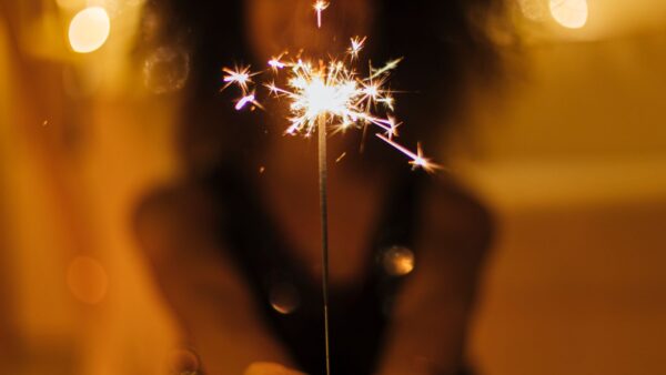 Pessoa segura uma vela faísca brilhante em destaque, com luzes desfocadas ao fundo, simbolizando celebração, esperança e um novo começo.