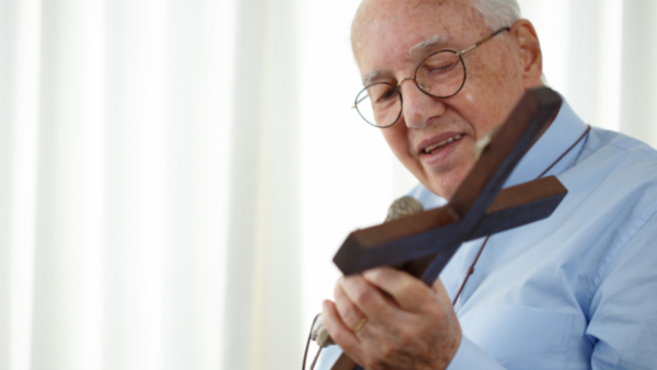 Monsenhor Jonas Abib de óculos e camisa azul clara segura um crucifixo de madeira com as duas mãos, olhando para ele com expressão serena e devota.