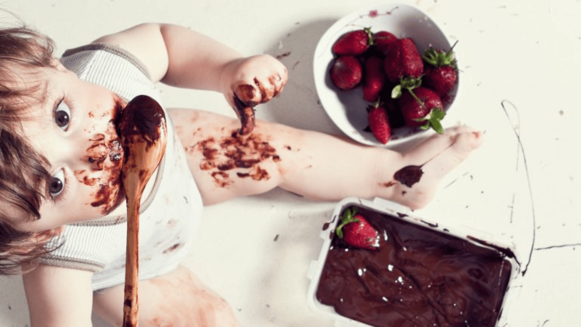 alt="Visão aérea de um bebê lambendo uma colher de pau suja de chocolate. Ele está sentado no chão, com o rosto, pernas e braços manchados de chocolate. Ao lado dele, há uma tigela com morangos frescos e um pote retangular cheio de calda ou creme de chocolate com um morango por cima."