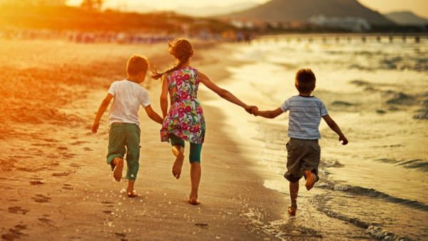 Férias sem telas: o que fazer para criar memórias reais com os filhos - Três crianças correm felizes e de mãos dadas pela beira-mar durante um pôr do sol dourado. A imagem transmite alegria e liberdade, com a luz do sol poente criando um brilho quente na areia e na água.