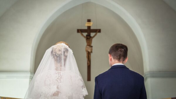 Visão traseira de um noivo e uma noiva durante uma cerimônia de casamento em uma igreja católica. À esquerda, a noiva usa um vestido de renda branca e um véu transparente. À direita, o noivo veste um terno azul-marinho. Ambos estão voltados para o altar, onde um crucifixo de madeira com a imagem de Jesus Cristo está fixado sob um arco arquitetônico claro ao fundo.