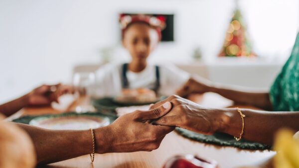 Família reunida à mesa durante uma refeição, com as mãos dadas em oração antes da refeição.