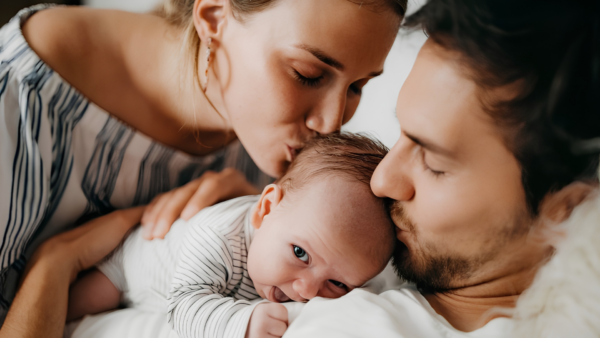 Fotografia em close de uma família demonstrando afeto. Um pai e uma mãe, ambos de olhos fechados, beijam carinhosamente a cabeça de seu bebê, que está deitado entre eles com os olhos abertos e uma expressão tranquila. A mãe veste uma blusa branca com listras escuras, o bebê usa uma roupinha clara listrada e o pai veste uma camiseta branca. O clima transmitido é de amor, união e acolhimento familiar.