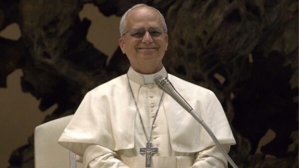 Papa Leão XIV sorri durante discurso ao microfone, vestido de branco e usando o crucifixo.
