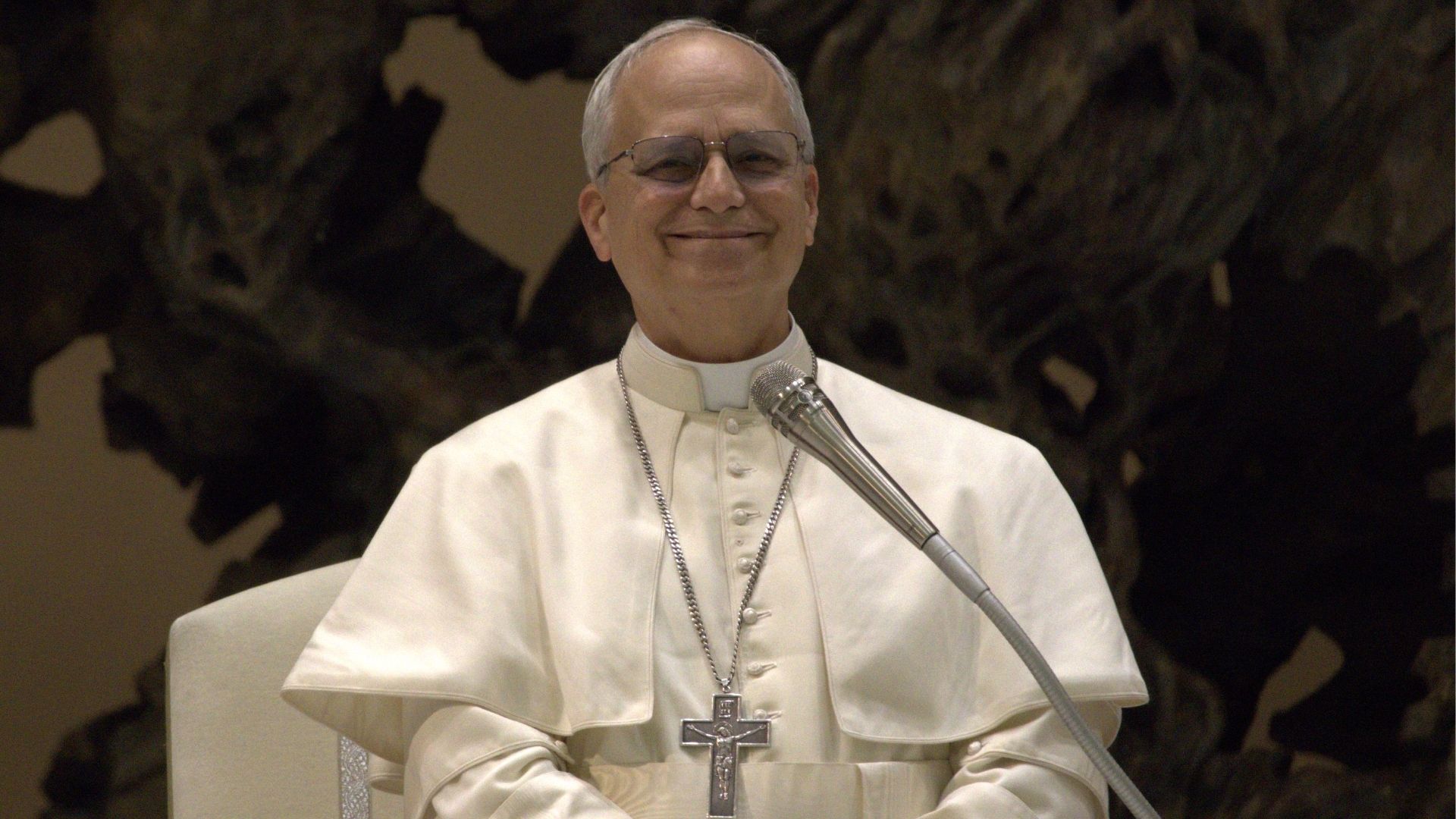 Papa Leão XIV sorri durante discurso ao microfone, vestido de branco e usando o crucifixo.