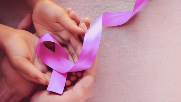 Mãos seguram uma fita rosa, símbolo da conscientização e prevenção do câncer de mama, sobre uma superfície de madeira clara.