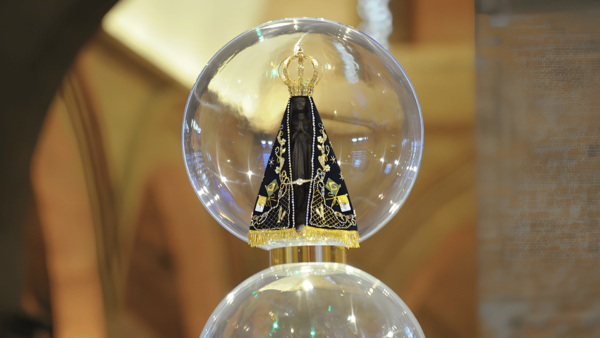 Fotografia em plano médio da imagem de Nossa Senhora Aparecida protegida dentro de uma cúpula transparente de vidro em formato esférico. A pequena estátua negra veste manto azul-marinho com bordados dourados, franjas e as bandeiras do Brasil e do Vaticano. Sobre a cabeça, ostenta uma coroa dourada trabalhada. O fundo desfocado exibe tons quentes e dourados do interior do Santuário Nacional.