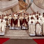 Um grande grupo de sacerdotes posa nos degraus do presbitério do Santuário do Pai das Misericórdias, todos vestidos com paramentos litúrgicos brancos e dourados. Ao centro, três novos sacerdotes usam casulas com detalhes em vermelho e dourado, ladeados por outros presbíteros. Atrás deles, destaca-se o bispo com mitra e báculo. A cena transmite solenidade e alegria após a celebração da ordenação presbiteral.