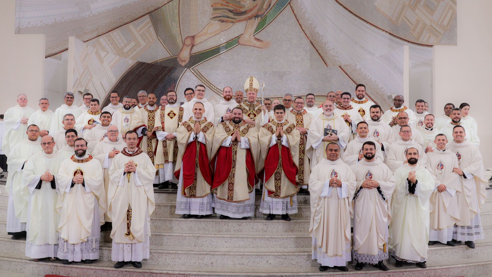 Um grande grupo de sacerdotes posa nos degraus do presbitério do Santuário do Pai das Misericórdias, todos vestidos com paramentos litúrgicos brancos e dourados. Ao centro, três novos sacerdotes usam casulas com detalhes em vermelho e dourado, ladeados por outros presbíteros. Atrás deles, destaca-se o bispo com mitra e báculo. A cena transmite solenidade e alegria após a celebração da ordenação presbiteral.